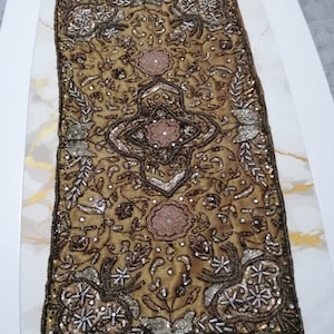 Könnte beinhalten: Ein goldener Tischläufer mit aufwendiger Stickerei, die ein Blumenmuster und silberne und braune Perlen zeigt.