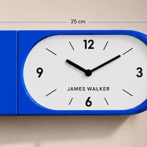 Op de afbeelding: Een rechthoekige blauwe wandklok met een afgeronde rechterkant, een witte wijzerplaat, zwarte wijzers en de zwarte cijfers 3, 6, 9 en 12. De tekst JAMES WALKER staat op de wijzerplaat. Afmetingen: 25 cm breed en 13 cm hoog.
