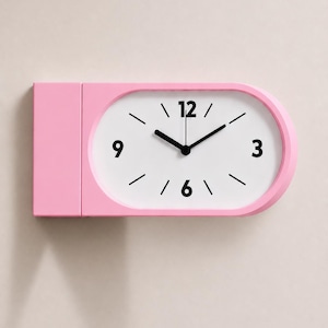 May include: A horizontal rectangular clock with a pink frame and a white face. The clock features black hour and minute hands, black numerals at the 3, 6, 9, and 12 positions, and simple black tick marks for the remaining hours.