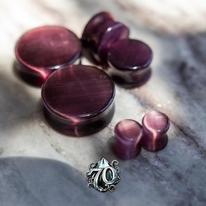 May include: A collection of five circular ear plugs in varying sizes, crafted from a deep purple material with a chatoyant cat's eye effect. The pieces feature a smooth, polished finish and a concave profile. A logo with the number 70 is visible.