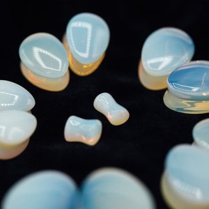 May include: A collection of iridescent opal ear gauges in various sizes and shapes, including teardrop, oval, and round. The gauges are made of a translucent, milky white material with a slight shimmer.