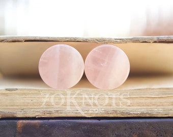 May include: Two round pink rose quartz plugs, each with a white stripe, are sitting on top of a closed book.