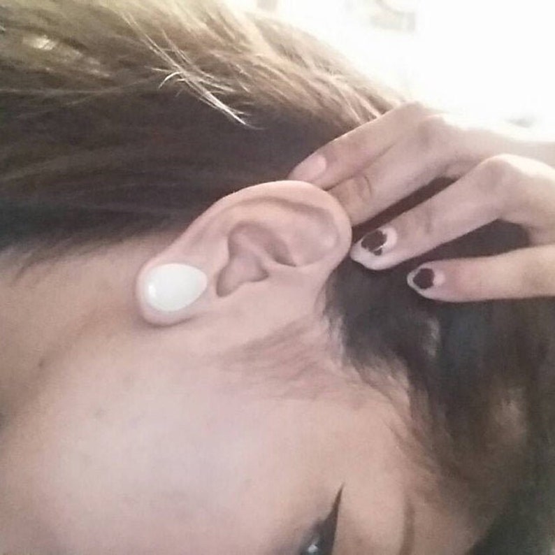 Puede incluir: Primer plano de la oreja de una persona con un expansor de oreja blanco. La persona tiene el pelo oscuro y lleva esmalte de u&ntilde;as negro.