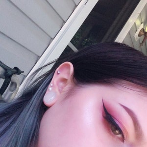 Puede incluir: Primer plano de la cara de una persona con cabello oscuro y un look de maquillaje de ojos dram&aacute;tico. La persona tiene m&uacute;ltiples piercings en la oreja, incluyendo un piercing redondo transparente de estilo tap&oacute;n.