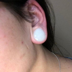 Puede incluir: Primer plano de la oreja de una persona con un tap&oacute;n para la oreja de vidrio redondo, liso y blanco.
