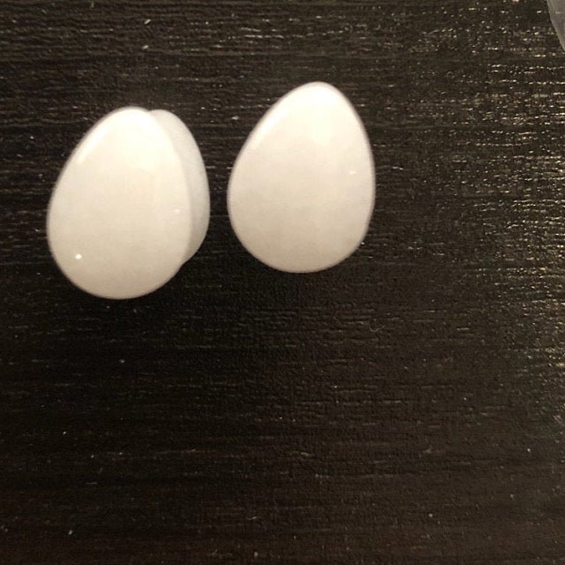 Puede incluir: Dos expansores de oreja blancos en forma de huevo, tambi&eacute;n conocidos como tapones para los o&iacute;dos o t&uacute;neles, sobre una superficie negra.