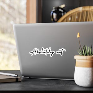 May include: A silver laptop computer with a black sticker that says "Absolutely not" on the lid. The laptop is sitting on a dark brown surface with a potted plant and a pen and notebook to the left.