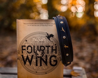 May include: A copy of the book Fourth Wing by Rebecca Yarros with a tan cover featuring black text and dragon illustrations, positioned next to a black leather-like strap decorated with gold-colored dragon silhouettes and small clear circular studs.