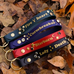 May include: Four leather keychains with gold lettering and rhinestones. The keychains are labeled "Fire heart", "Cursebreaker", "Starborn", and "Lady Death".