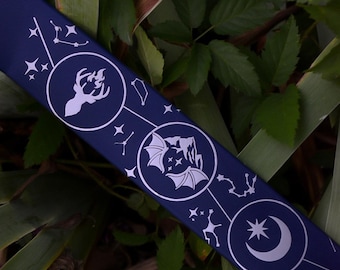 May include: A dark blue ribbon featuring a white printed design of three circular icons. The icons depict a deer head, bat wings over mountains, and a crescent moon with a star. Constellations and small stars connect the circular motifs.