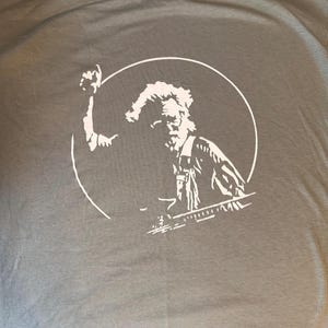 May include: A gray t-shirt featuring a white graphic of a man within a white circle. The man is depicted with a raised arm and a beard, suggesting a vintage or folk art style. The shirt's fabric appears soft.