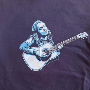 May include: A blue and white graphic print of a person playing an acoustic guitar on a dark blue t-shirt.