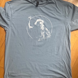 K&ouml;nnte beinhalten: Ein hellblaues T-Shirt mit einer wei&szlig;en Grafik einer Person, die ein Musikinstrument spielt. Die Grafik befindet sich in einem Kreis.