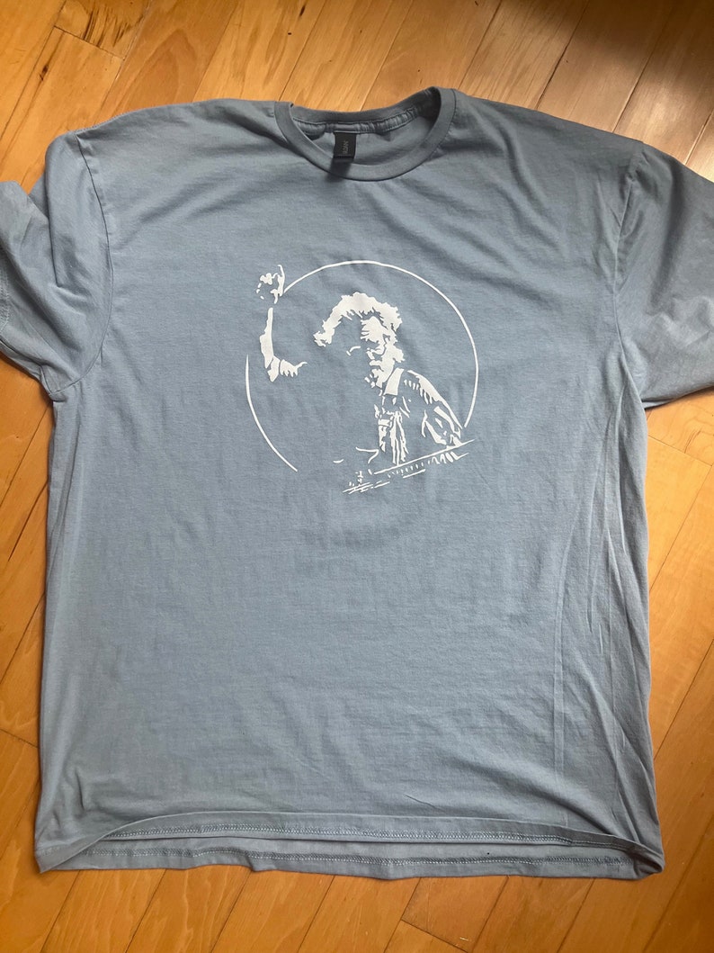 K&ouml;nnte beinhalten: Ein hellblaues T-Shirt mit einer wei&szlig;en Grafik einer Person, die ein Musikinstrument spielt. Die Grafik befindet sich in einem Kreis.