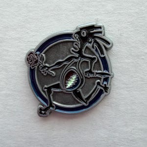 May include: A silver-toned pin featuring a stylized skeleton figure with a rose and a lightning bolt design on its belly. The figure is set against a blue-ringed circle. The pin has a detailed, artistic design.