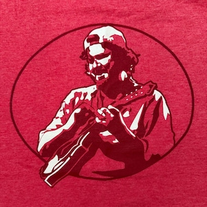May include: Red t-shirt featuring a graphic of a person in a baseball cap, within a circular frame. The graphic is in shades of red and white, depicting the person seated with their legs drawn up.