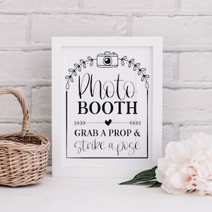 May include: White sign with black text that reads "Photo Booth Grab a Prop & Strike a Pose". A camera icon is above the text. The sign is on a white brick wall.