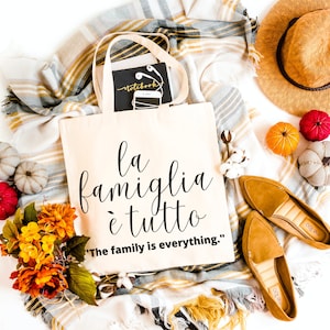May include: A beige tote bag with the text "la famiglia &egrave; tutto" in black cursive script. The text is translated below in English as "The family is everything."