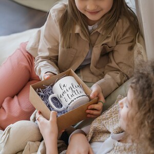 May include: Two children are sitting on a bed, one is giving the other a gift box containing a white mug with the text "La famiglia &egrave; tutto" and the words "Family is everything" in smaller font below.