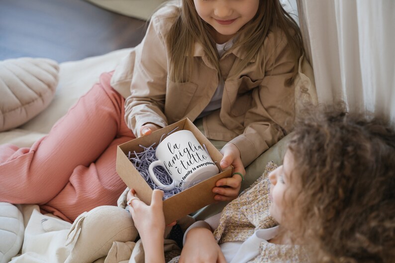 May include: Two children are sitting on a bed, one is giving the other a gift box containing a white mug with the text "La famiglia &egrave; tutto" and the words "Family is everything" in smaller font below.