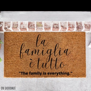 May include: A brown coir doormat with the text "la famiglia &egrave; tutto" in black cursive script. The text is translated below in English as "The family is everything."