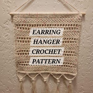 May include: A cream-coloured crochet earring hanger with a wooden dowel and hanging string. The text "EARRING HANGER CROCHET PATTERN" is displayed on the front. The bottom edge features several tassels with pink accents.