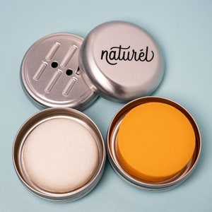 Op de afbeelding: Drie zilveren metalen containers met deksels. E&eacute;n container heeft een label met de tekst "naturel". E&eacute;n container bevat een witte zeepstaaf. E&eacute;n container bevat een gele zeepstaaf.