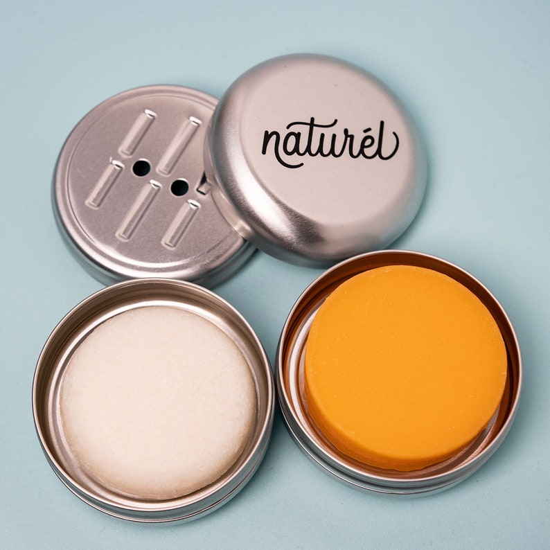 Op de afbeelding: Drie zilveren metalen containers met deksels. E&eacute;n container heeft een label met de tekst "naturel". E&eacute;n container bevat een witte zeepstaaf. E&eacute;n container bevat een gele zeepstaaf.