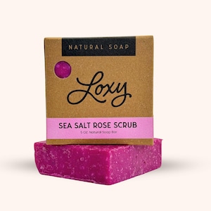 May include: A pink sea salt rose scrub soap bar with the text "Loxy" and "SEA SALT ROSE SCRUB 5 OZ. Natural Soap Bar" on the packaging.