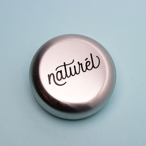 Op de afbeelding: Een ronde zilveren container met het woord "naturel" in zwarte cursieve letters.