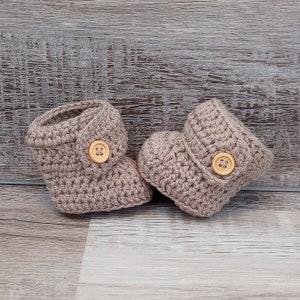 May include: A pair of crocheted baby booties in a light brown color with a button on each bootie.