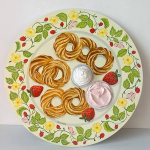 May include: A white ceramic plate with a green floral border featuring yellow flowers and red strawberries. The plate has a variety of pastries, including a ring-shaped pastry, and two small bowls of white and pink frosting.