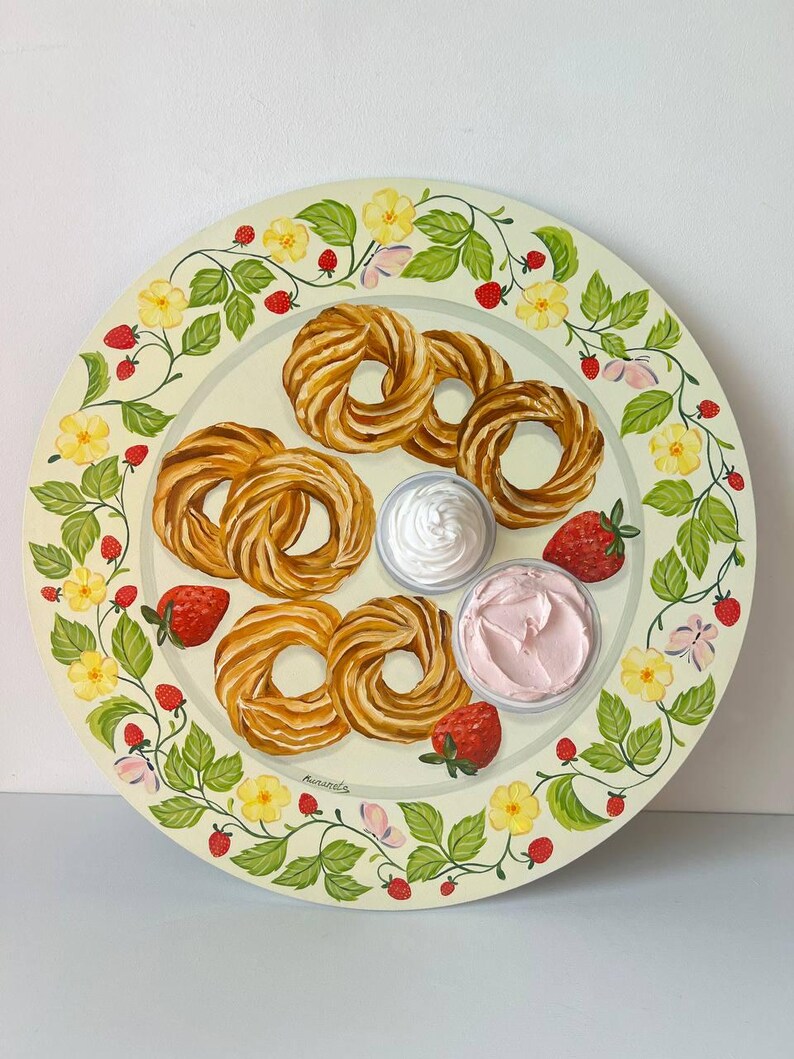 May include: A white ceramic plate with a green floral border featuring yellow flowers and red strawberries. The plate has a variety of pastries, including a ring-shaped pastry, and two small bowls of white and pink frosting.