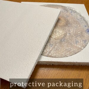 May include: A white styrofoam box with a lid, protecting a round, clear glass plate with a blue and white pattern. The box is sitting on a wooden surface.  The text "protective packaging" is printed on the surface.