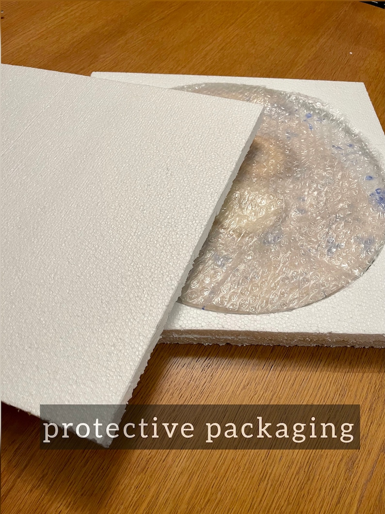 May include: A white styrofoam box with a lid, protecting a round, clear glass plate with a blue and white pattern. The box is sitting on a wooden surface.  The text "protective packaging" is printed on the surface.