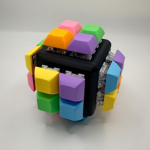 以下が含まれることがあります： 黒い中央コアの各面にメカニカルキーボードスイッチが取り付けられた立方体型のフィジェットトイ。スイッチの上部には、ピンク、オレンジ、緑、紫、黄色、青のマットな質感のキーキャップが装着されています。