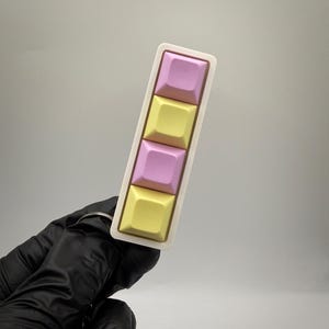 May include: A rectangular keychain featuring four mechanical keyboard keycaps arranged in a vertical row. The keycaps alternate between pastel pink and pastel yellow colors, all set within a white frame with a metal ring attached at the base.