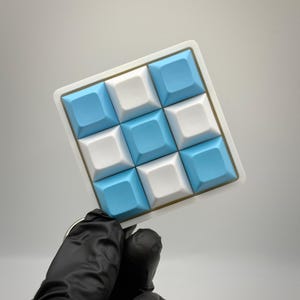 May include: A square keychain featuring a three-by-three grid of nine mechanical keyboard keycaps arranged in a checkerboard pattern. The keycaps alternate between matte white and light blue colors, all mounted on a white square base.