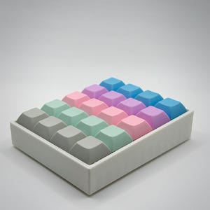 May include: A rectangular tray filled with colorful, square keyboard keycaps. The keycaps are arranged in rows and columns, featuring a gradient of colors including gray, mint green, pink, purple, and blue. The tray is white, and the background is a plain white.