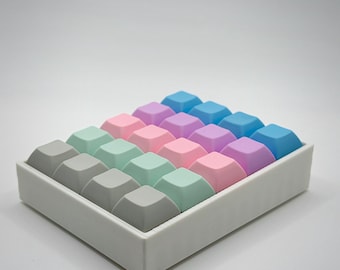May include: A rectangular white tray filled with colorful, square-shaped keycaps. The keycaps are arranged in rows and columns, with a gradient of colors including gray, mint green, pink, purple, and blue. The background is a plain white.
