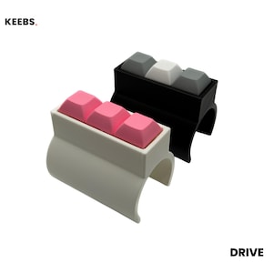 May include: Two mechanical keyboard switch rings labeled KEEBS. and DRIVE. One ring is white with three pink keycaps, and the other is black with two gray keycaps and one white keycap in the center. Each ring features a curved, open-ended band.