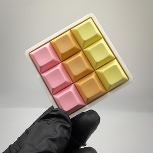 May include: A square-shaped keypad accessory featuring a three-by-three grid of nine individual keys. The keys are arranged in a color gradient, transitioning from soft pink on the left to orange in the center and light yellow on the right side.