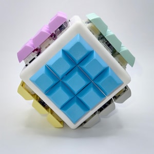 May include: A cube-shaped keyboard with a white frame and colorful keycaps. The keycaps are in shades of blue, pink, yellow, and green. The keyboard is designed with a unique, geometric aesthetic.