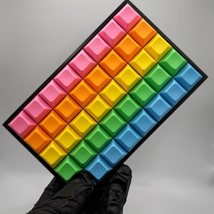 May include: A rectangular mechanical keyboard macro pad featuring a grid of forty-two square keycaps arranged in a rainbow gradient pattern, transitioning from pink and orange to yellow, green, and blue, set within a black frame.