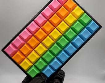 May include: A rectangular mechanical keyboard macro pad featuring a grid of forty-eight square keycaps arranged in a rainbow gradient pattern. The keycaps transition from pink to orange, yellow, green, and blue, all set within a black frame.