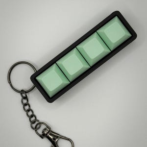 Könnte beinhalten: Ein rechteckiger schwarzer Schlüsselanhänger mit drei hellen mintgrünen, quadratischen mechanischen Tastaturtasten in einer Reihe. Der Schlüsselanhänger verfügt über einen dunklen Metallring und einen Karabinerhaken an einer kurzen Kette.