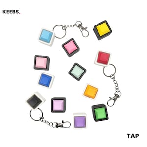 Op de afbeelding: Een verzameling kleurrijke vierkante sleutelhangers met vierkante knoppen in verschillende kleuren, waaronder blauw, roze, geel en groen. Elke sleutelhanger heeft een zilveren ketting en sluiting. Het woord "KEEBS" staat linksboven en het woord "TAP" rechtsonder.