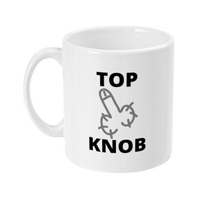May include: White ceramic mug featuring a black and grey graphic of a hand gesture and the words "TOP KNOB". The mug has a curved handle and a cylindrical shape.
