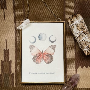 Könnte beinhalten: Ein gerahmter Druck mit einer Illustration eines Schmetterlings und Mondphasen, aufgehängt an einer goldenen Kette. Der Druck enthält den Text "It's natural to outgrow your old self." Ein klarer Quarzkristall und ein Salbeibündel sind daneben.