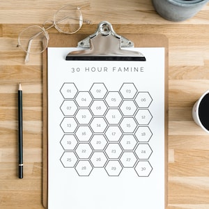 May include: A clipboard with a white paper featuring the text "30 HOUR FAMINE" and a grid of numbered hexagons. A pair of round glasses and a pencil are on the wooden surface. A potted plant and a cup of coffee are also visible.